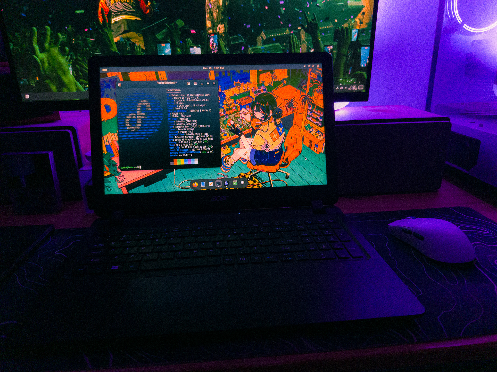 Laptop on a desk showing a Linux desktop with a terminal window running Fastfetch, with the Fedora logo on ASCII, and illustrated wallpaper, placed in front of an external monitor and next to a desktop PC in a dimly lit room with purple ambient lighting