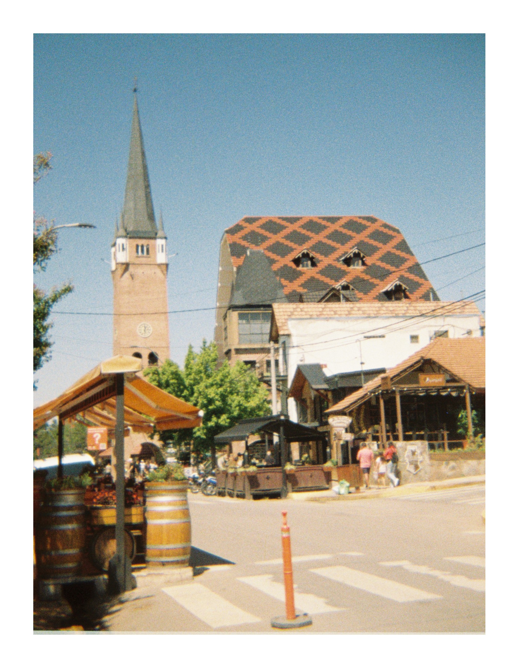 Street intersection in a small town with pedestrians crossing, outdoor seating areas, and buildings with sloped roofs, including a clock tower in the background under clear daylight.