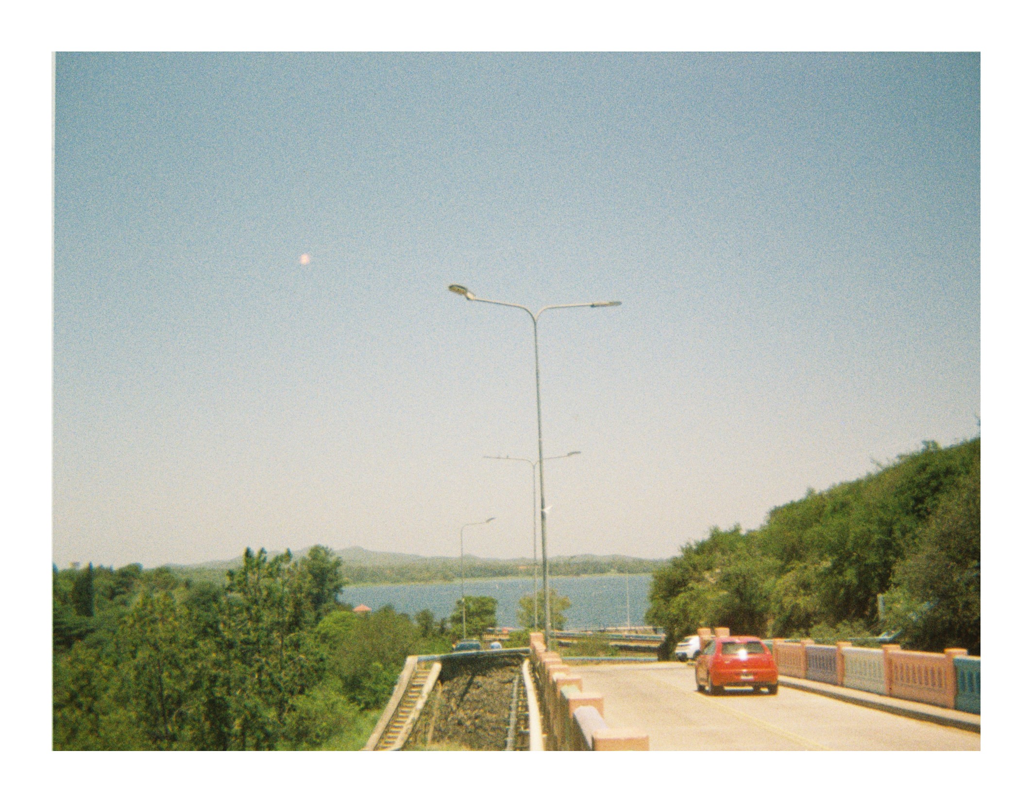 Road bridge with cars driving toward a lake in the distance, bordered by trees and streetlights under clear daylight."