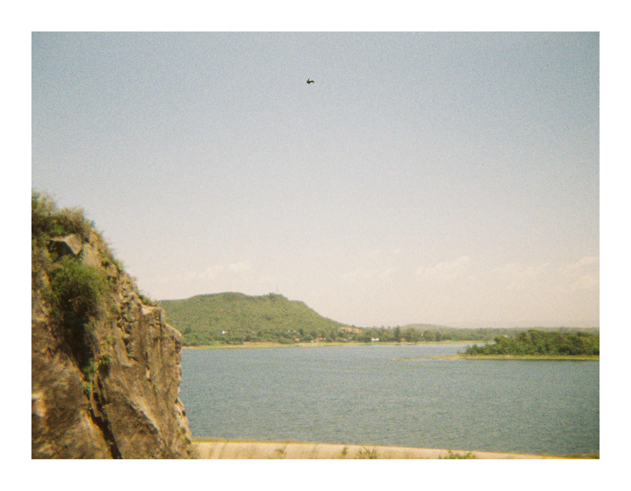 Rocky cliff in the foreground overlooking a wide lake, with the edge of a dam visible below and hills and shoreline in the distance under a pale sky.