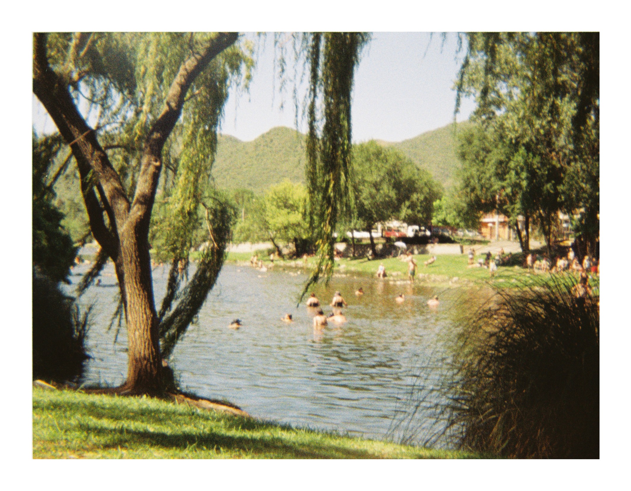 People swimming and standing in a shallow river, with trees along the shore and grassy banks, and hills visible in the background under daylight.