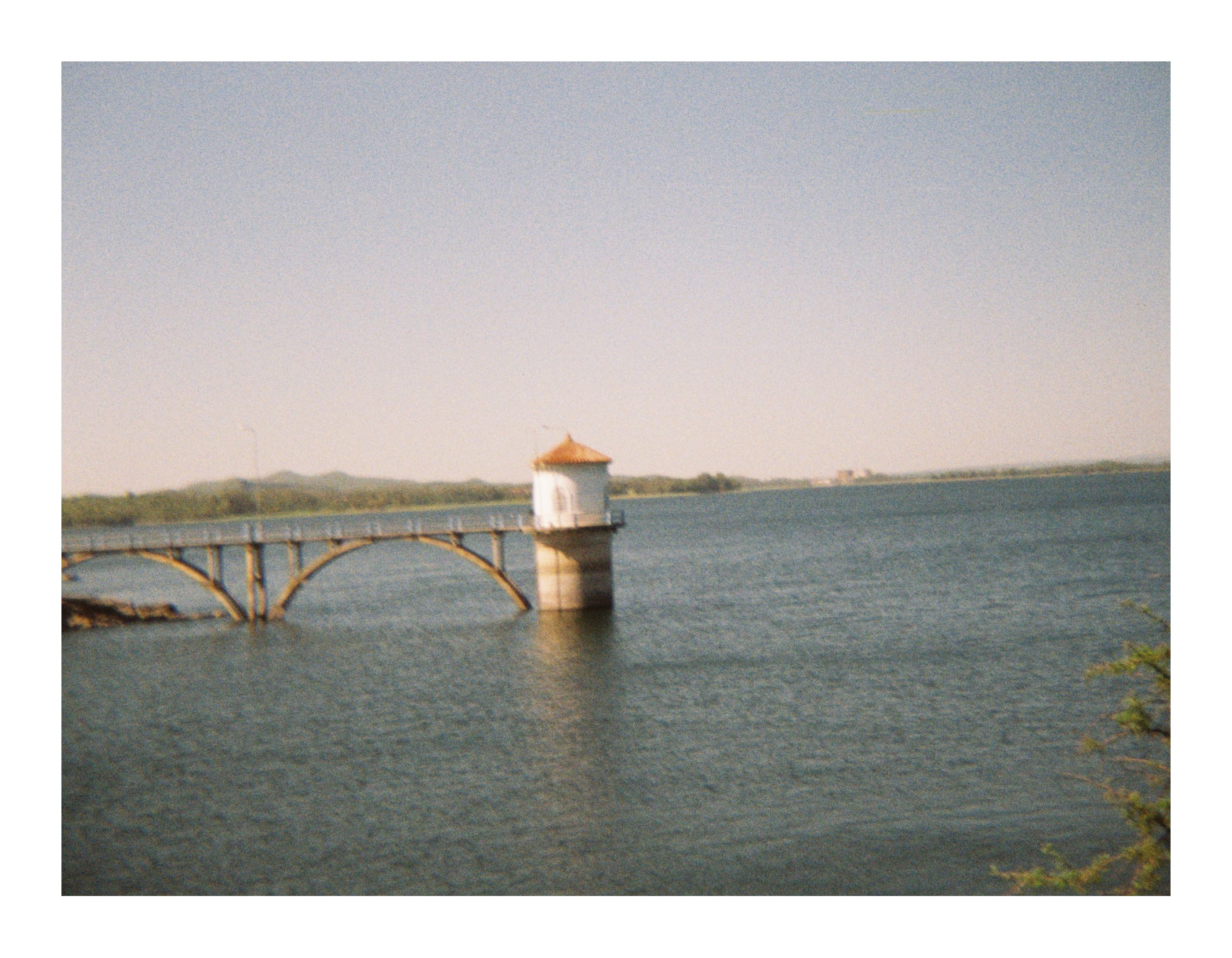 Small tower structure connected by a narrow bridge extending over a lake, with calm water and distant shoreline under clear daylight.