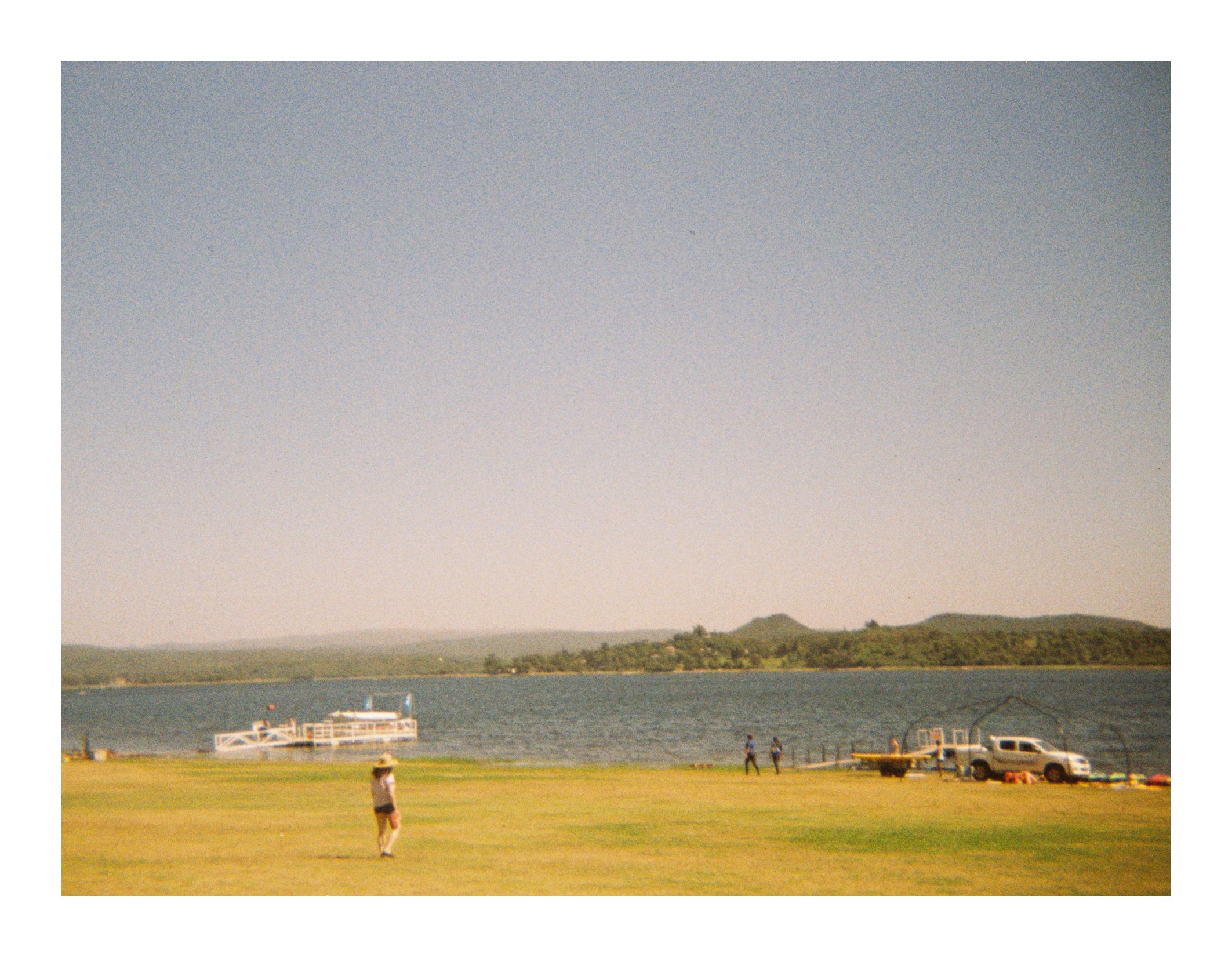 Open grassy area by a lake with a dock and boats, a few people walking near the water, and hills visible across the lake under clear daylight.